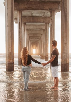 Couple holding hands while standing in shallow water under beach pier at sunset.