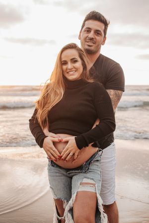 Sunset beach maternity photo with couple embracing under cloudy sky.