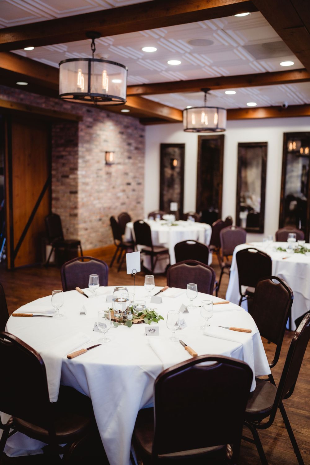 An elegant reception venue with round tables set with white linens and dark chairs under pendant lights.