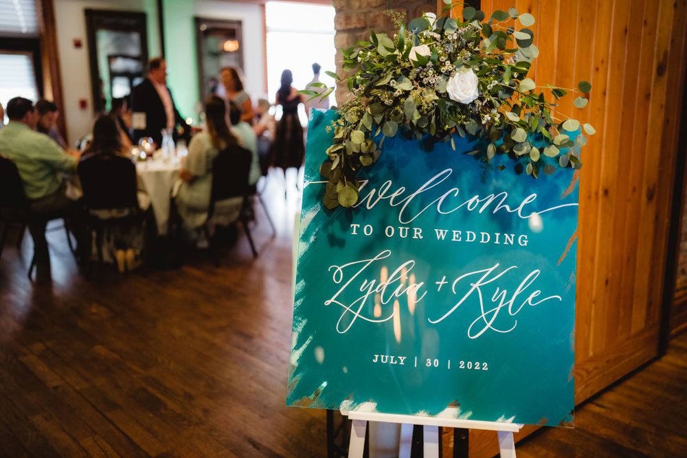 A teal wedding welcome sign with floral decorations displayed on a wooden easel.