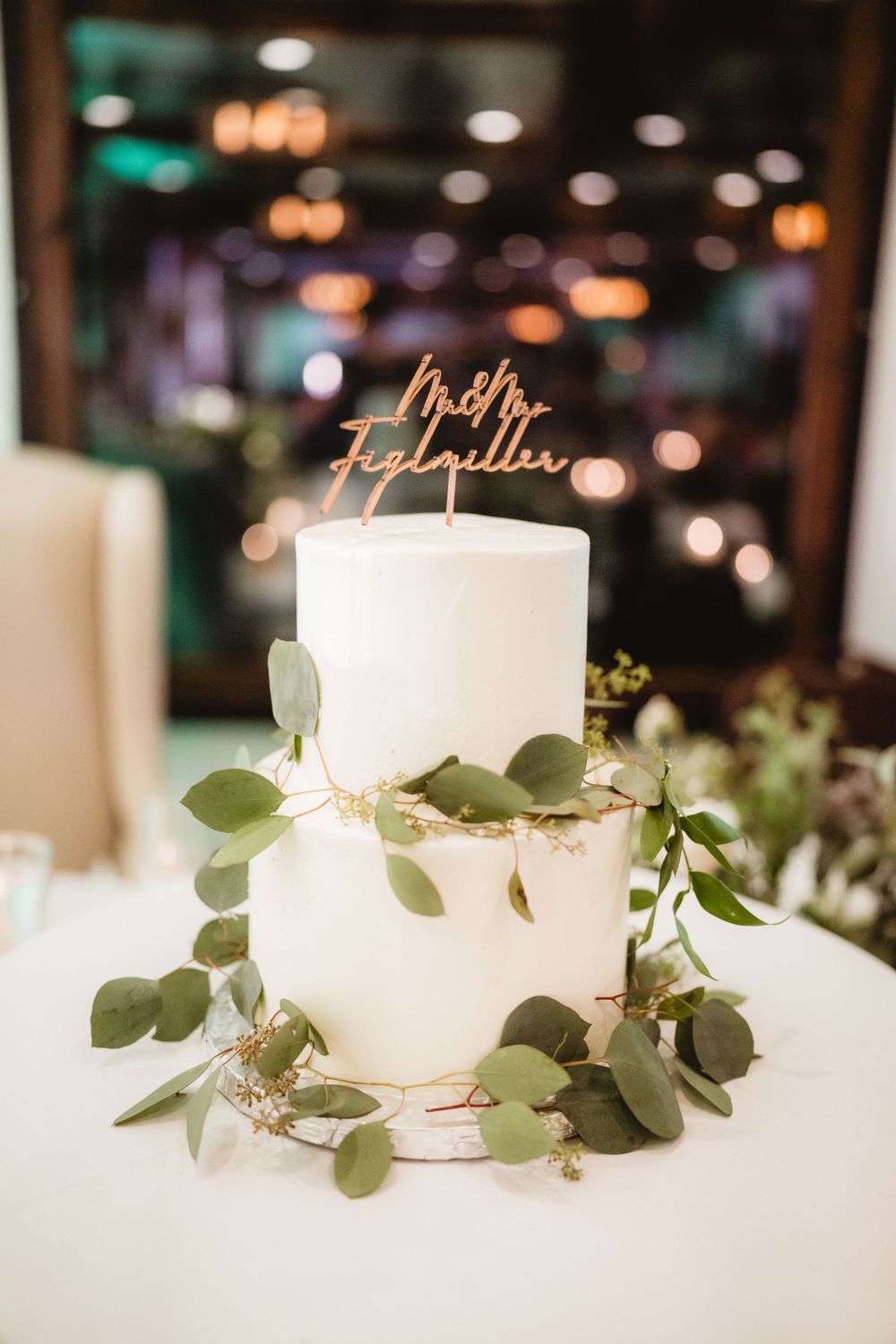 A simple white two-tier wedding cake decorated with eucalyptus leaves and a gold topper.