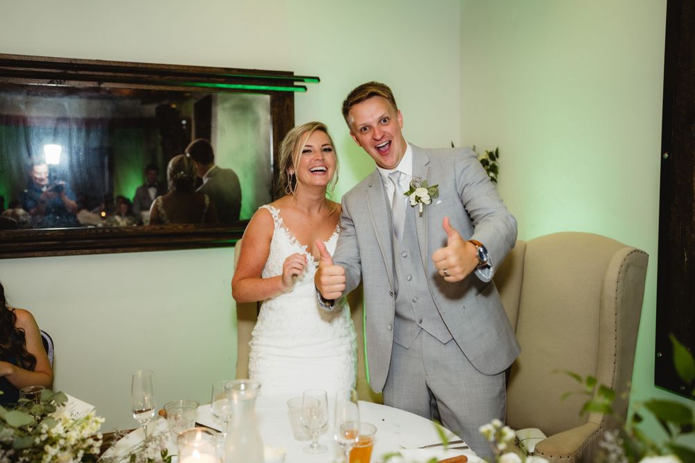 A newlywed couple gives thumbs up while celebrating at their reception table.