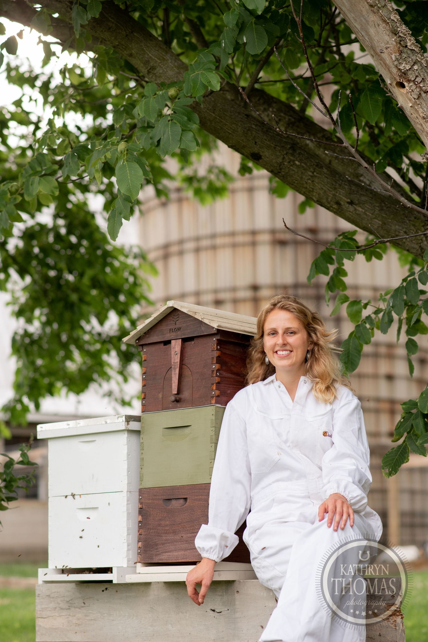 A beekeeper in white attire stands next to stacked wooden beehives under a leafy tree.