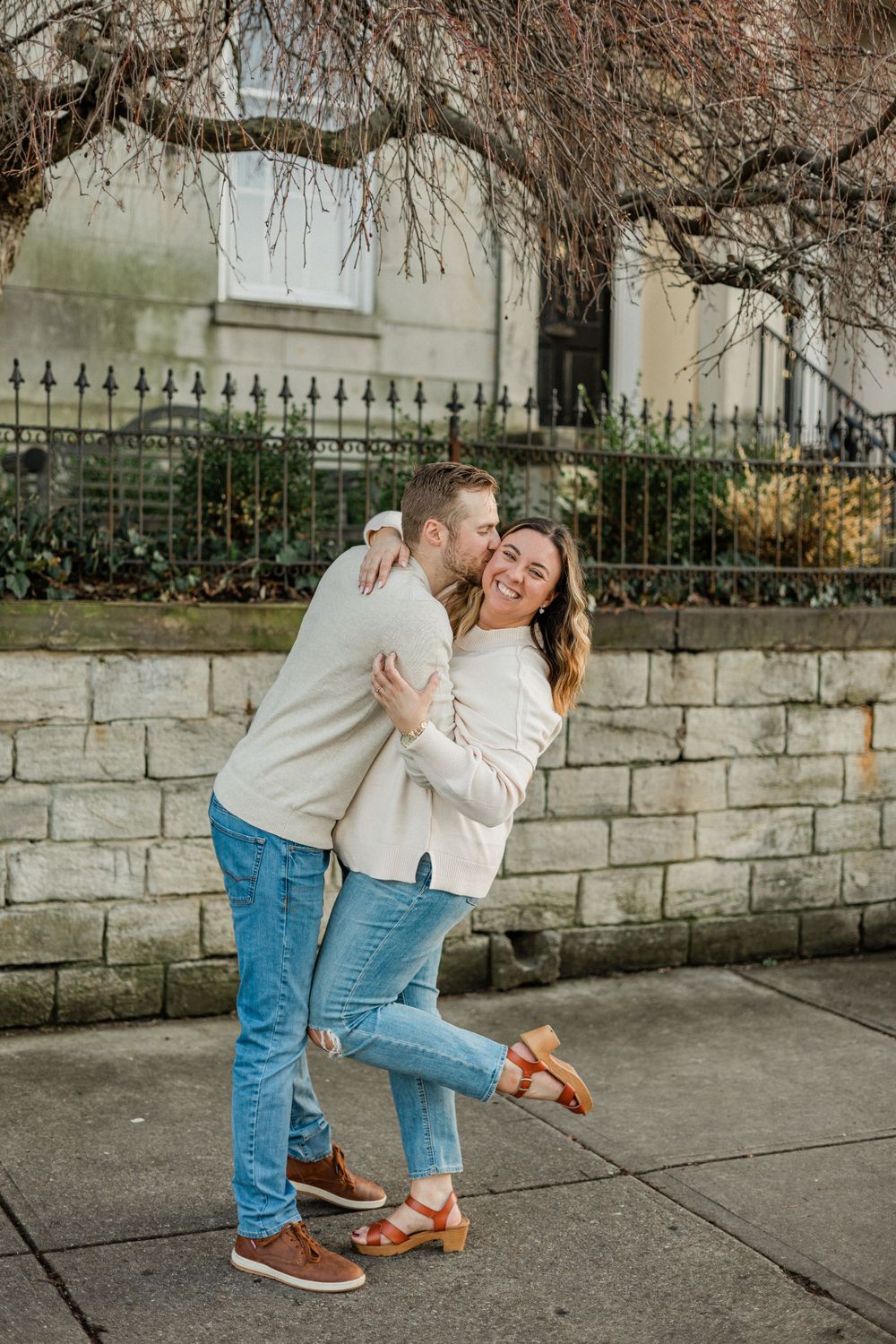 Joyful couple embracing and laughing against a stone wall with bare winter branches overhead.