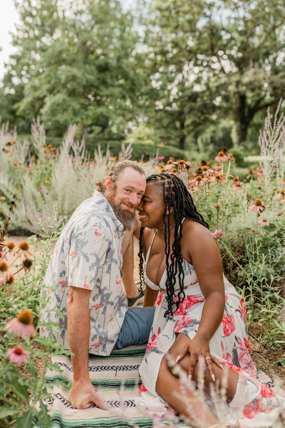 A couple laughs together while sitting on a blanket surrounded by wildflowers and greenery.