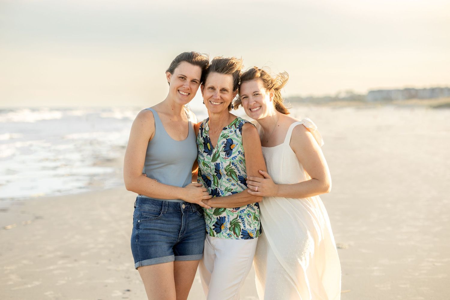 Three figures share a close moment together on a sandy beach with ocean waves in the background.