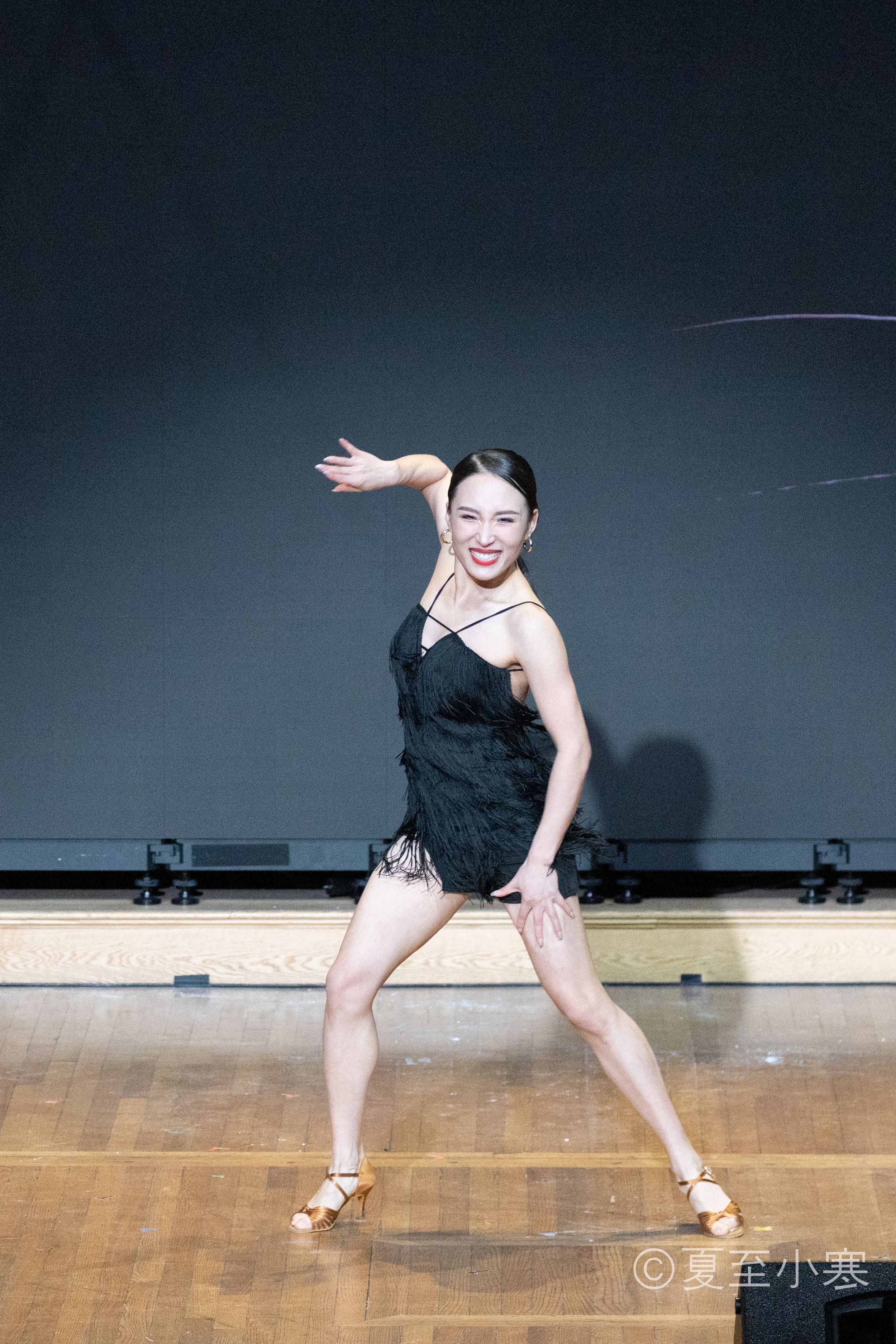 Dancer in black dress performs graceful stretching movements on wooden floor with dramatic lighting.