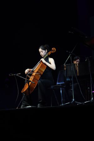 Solo cellist performing on a dimly lit concert stage with piano in background.