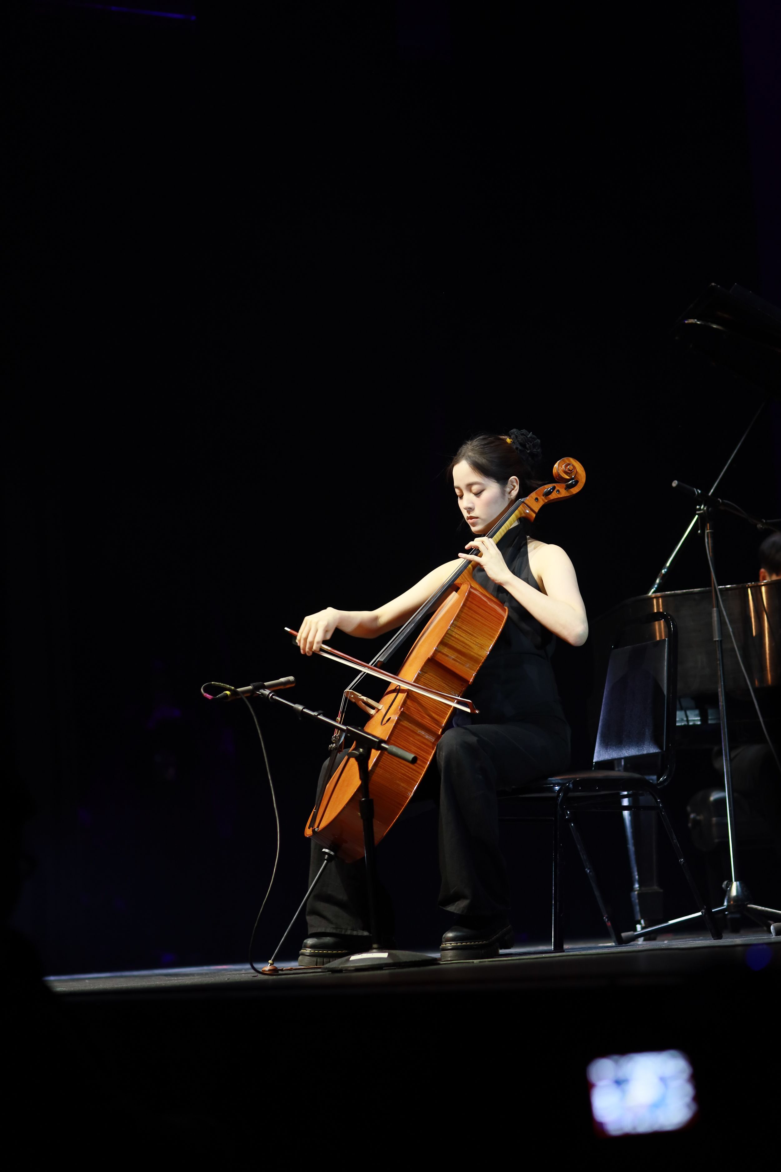Cellist performing on stage in a black dress against a dark background.