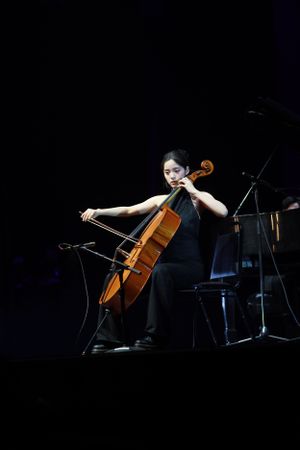 Dramatic stage performance of a cellist in black attire against dark backdrop.