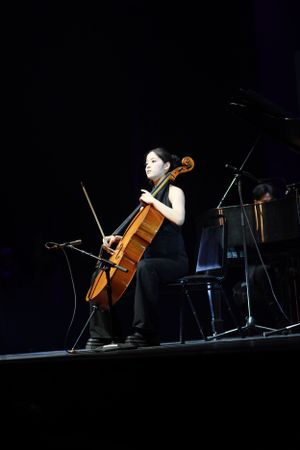 Cellist in black performing on stage with dramatic lighting.