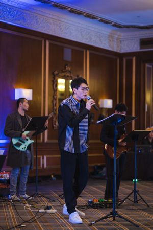 A performer sings into a microphone on stage while accompanied by a guitarist in an elegant ballroom setting.