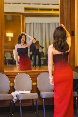 Woman in a red ombre dress checks her appearance in a large mirror surrounded by chairs.