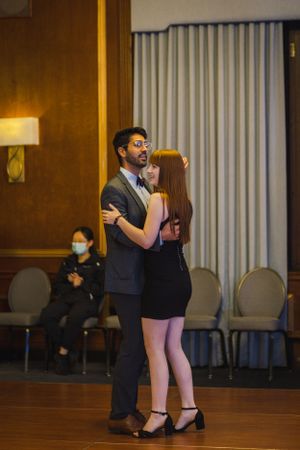 Dancing couple in formal wear practicing steps in ballroom with chairs in background.