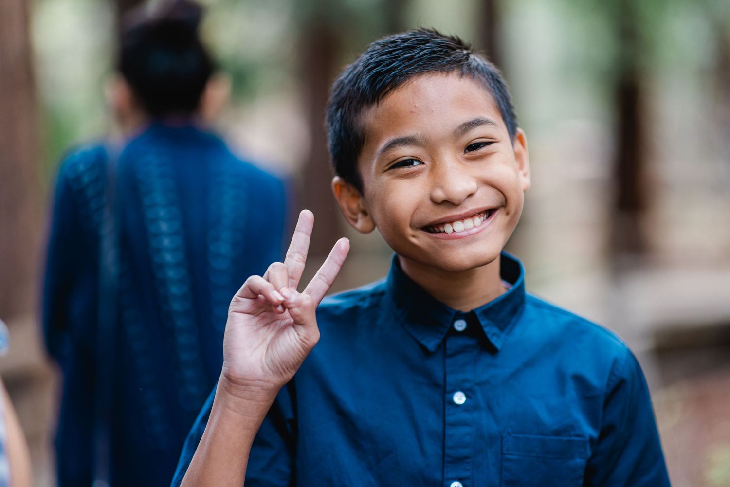 Young person in a navy blue polo shirt making peace sign while smiling in forest setting.