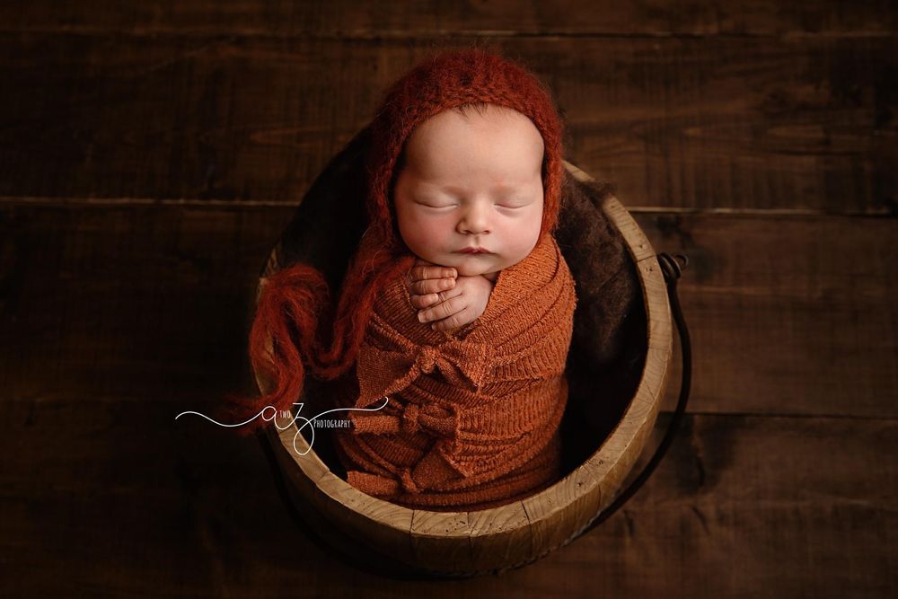Newborn photography featuring cozy orange wrap and rustic wooden barrel setting.