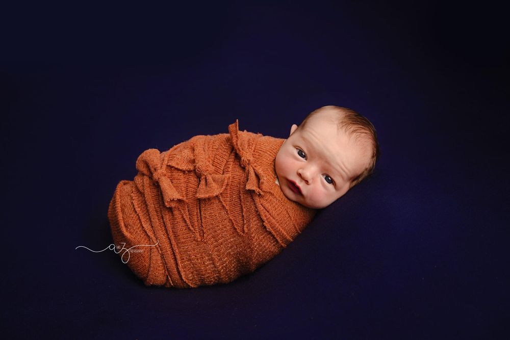 Newborn wrapped in orange knit blanket against dark blue background in professional photo setup.