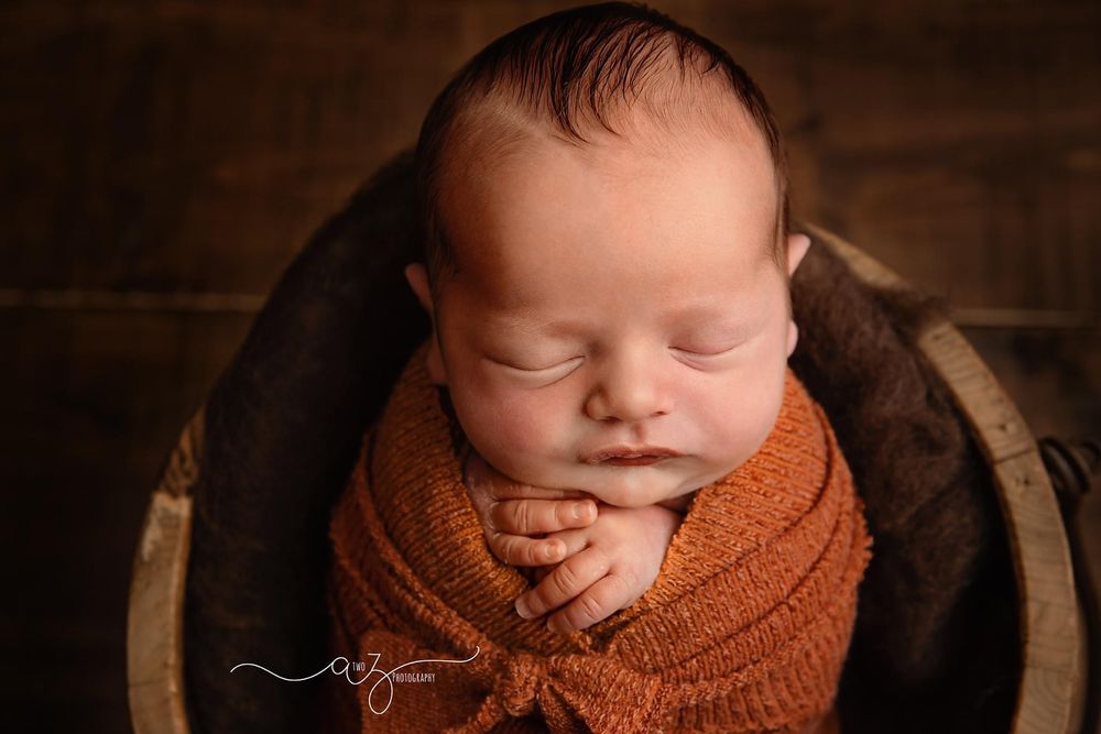 Close-up portrait of sleeping newborn swaddled in orange wrap against dark wooden background.