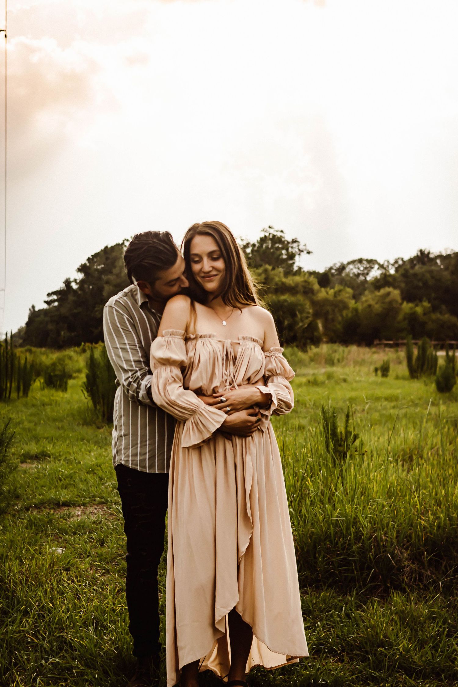 Romantic couple shares intimate moment in flowing beige dress against green field at sunset.