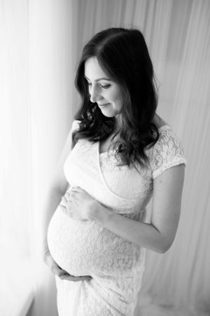Natural light maternity portrait of woman in white dress looking happily at her bump