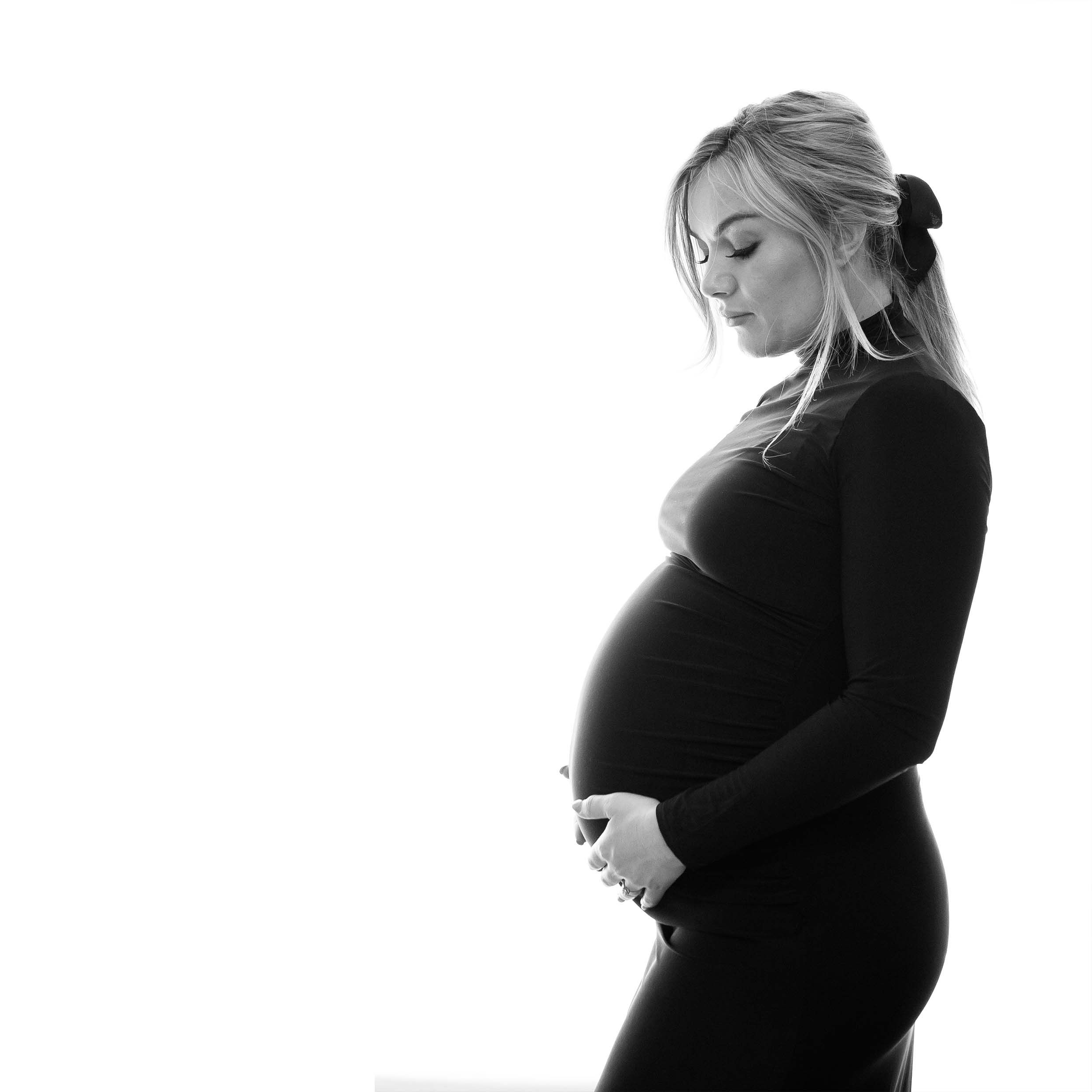 Back lit maternity portrait of blonde woman looking at her bump in profile