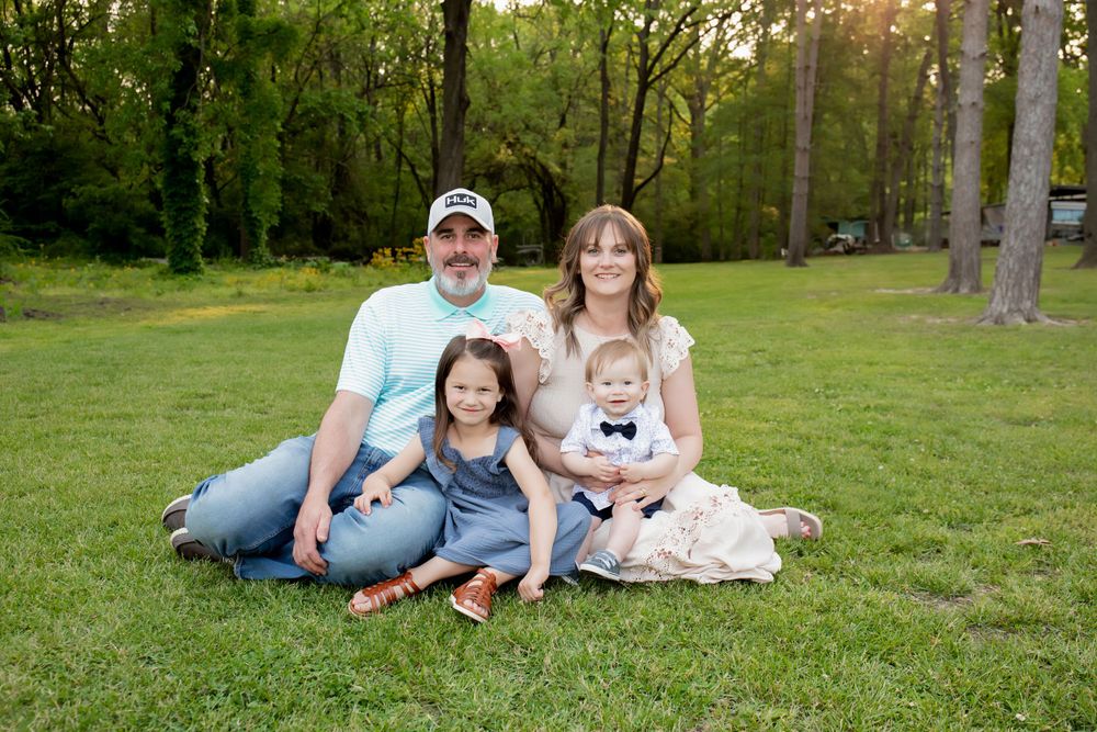 A family of four sits together on grass with a wooded background during golden hour.
