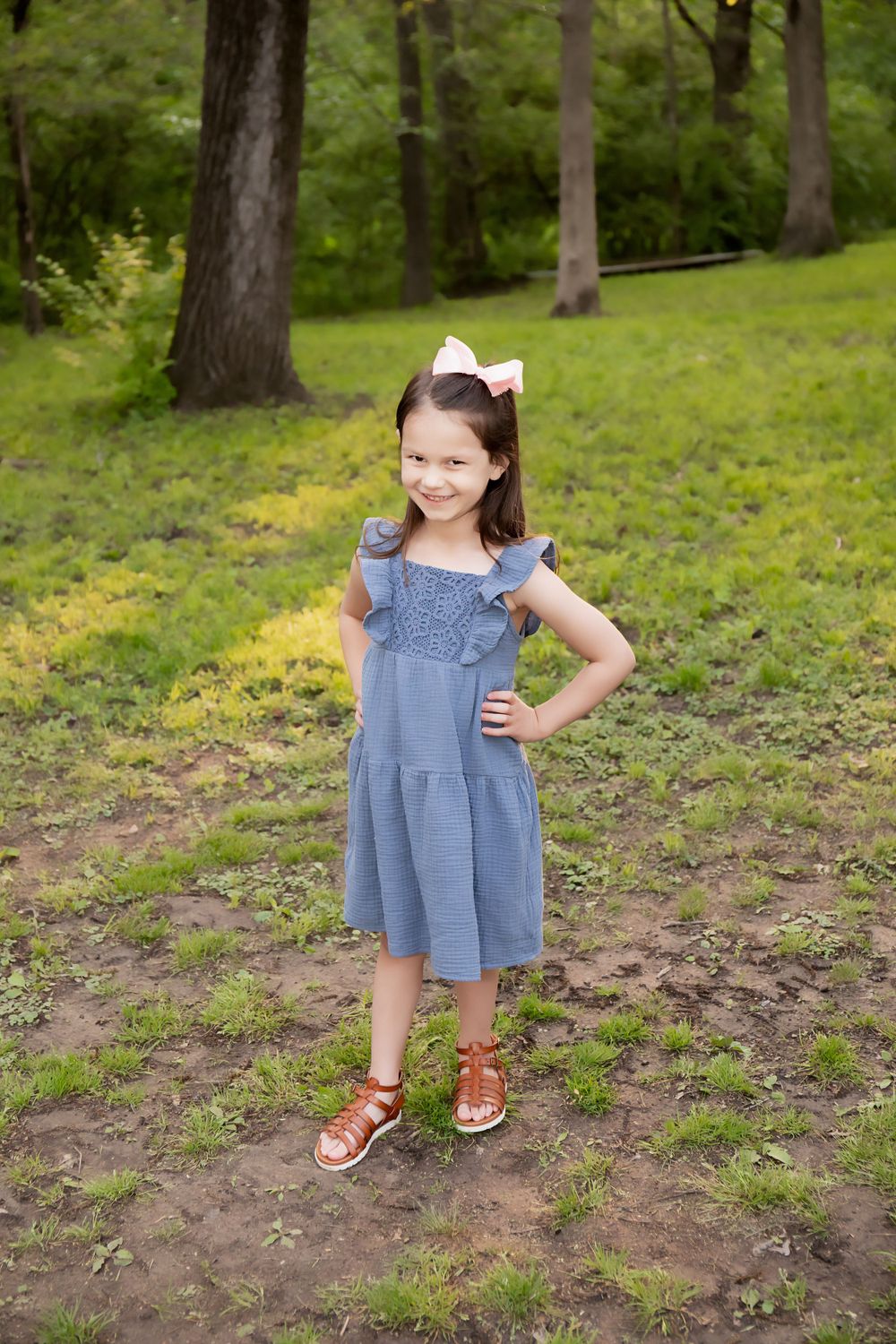 A child poses confidently in a blue dress and white bow against a forest backdrop.