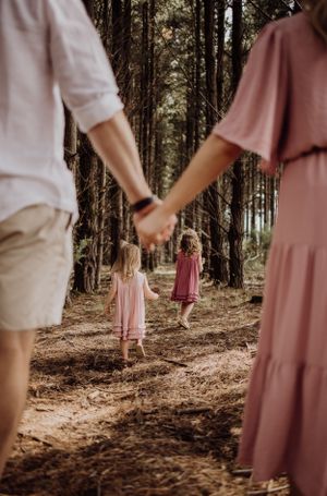 Parents holding hands while children in pink dresses walk through a pine forest.