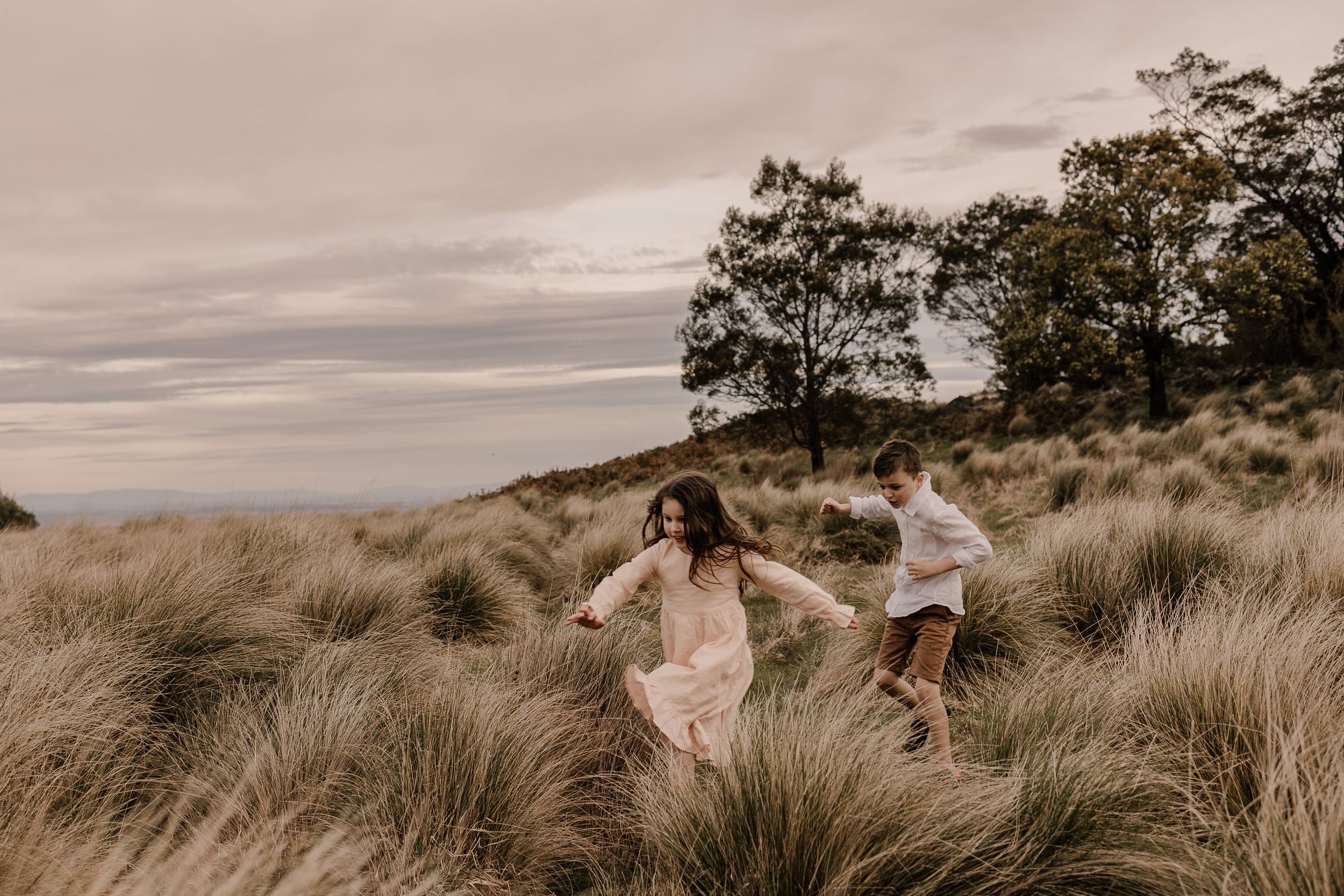 Two people running through tall grass on a hill during sunset.