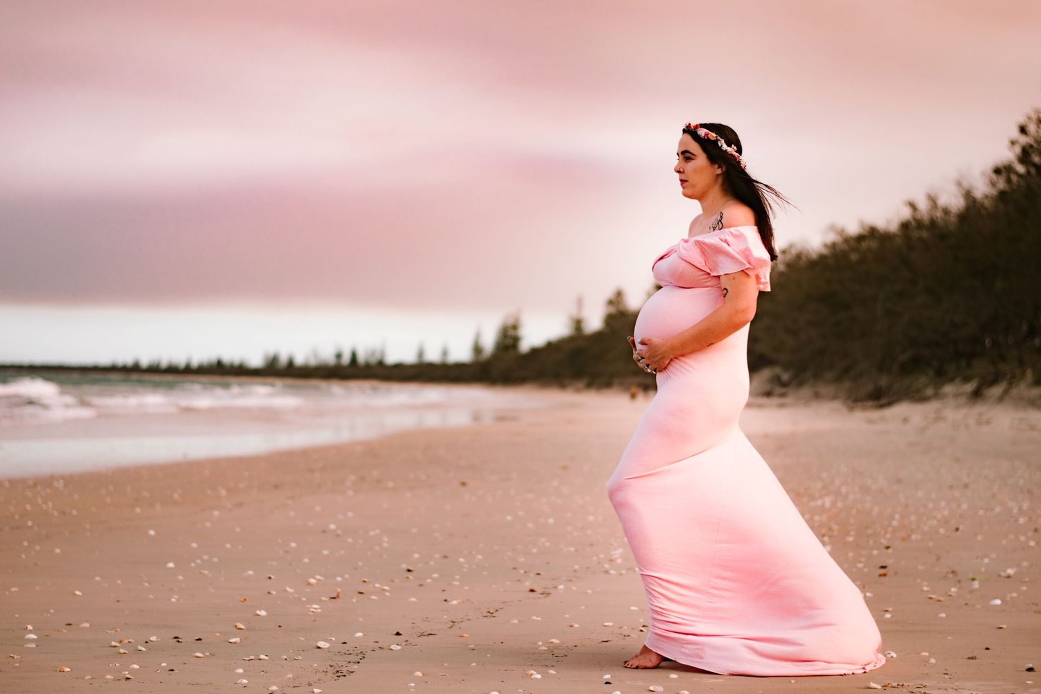 Pregnant woman in flowing pink dress standing on beach at sunset.