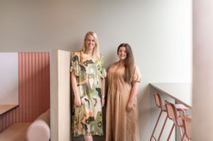 Two women standing together wearing floral print and neutral maxi dresses in a pink paneled interior space.