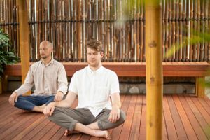 Two people practicing meditation on wooden deck with bamboo fence background.