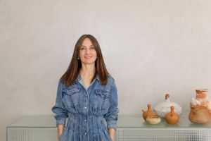 Woman wearing denim jumpsuit standing against neutral wall with ceramic vases.