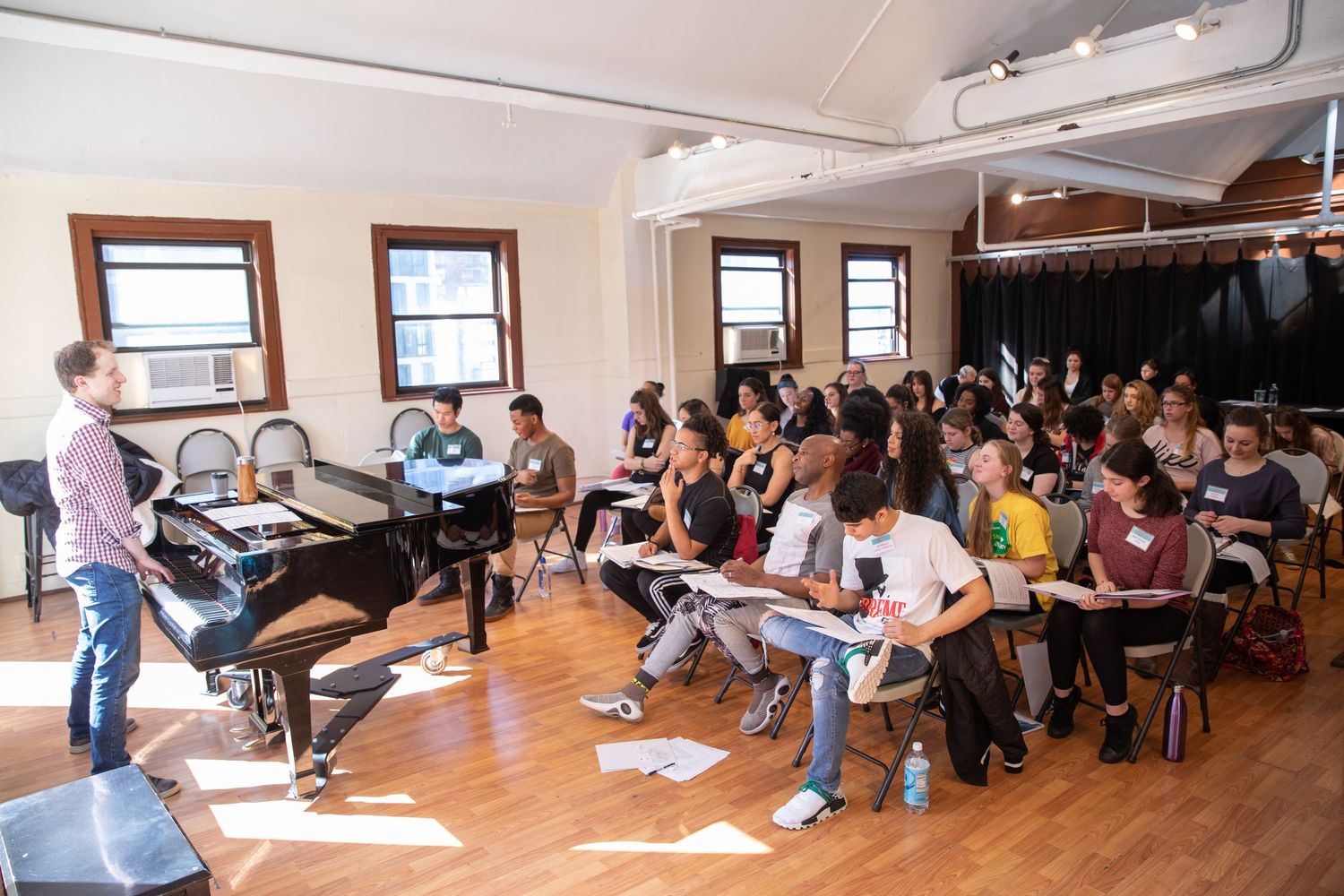 A music instructor leads a large group of students in a bright studio classroom with a grand piano nearby.