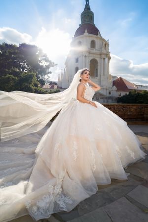 Bride in flowing white gown poses by historic church dome in dramatic sunlight.