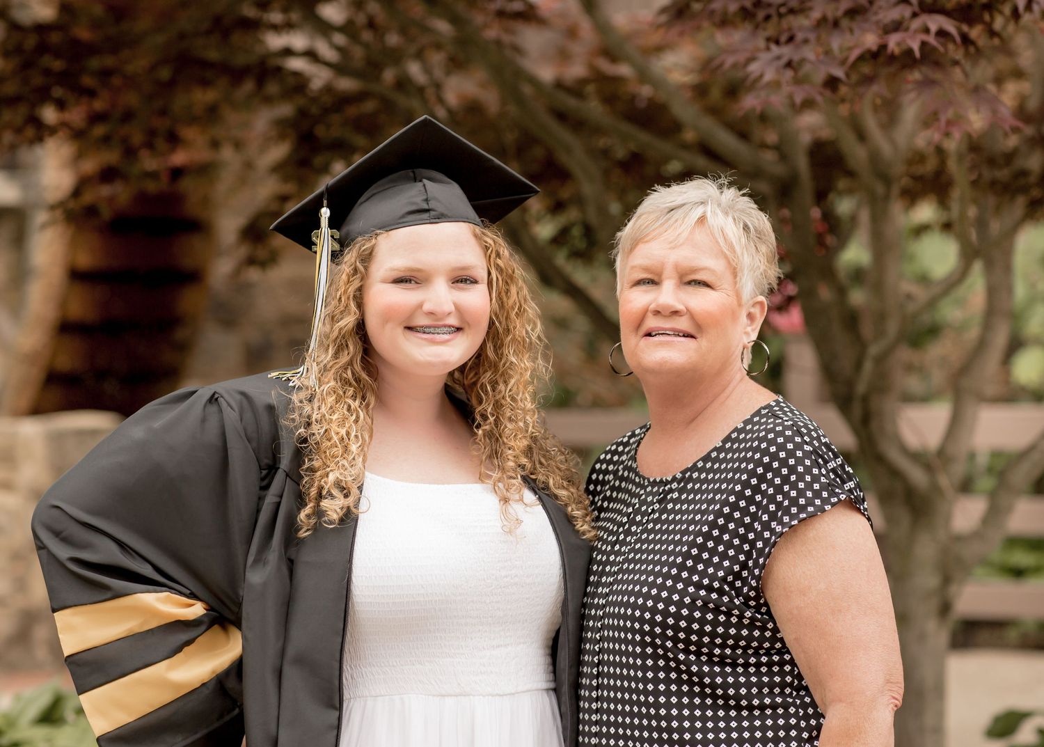 Graduate in cap and gown smiles with older woman at outdoor graduation ceremony.