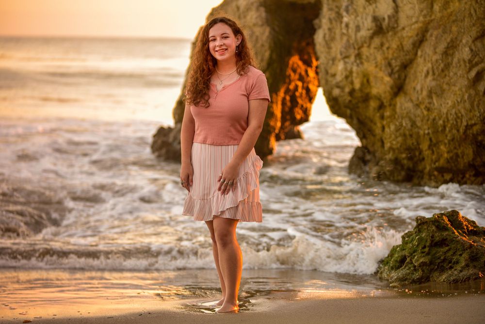 Woman in pink dress walking barefoot on beach at sunset near coastal cliffs.