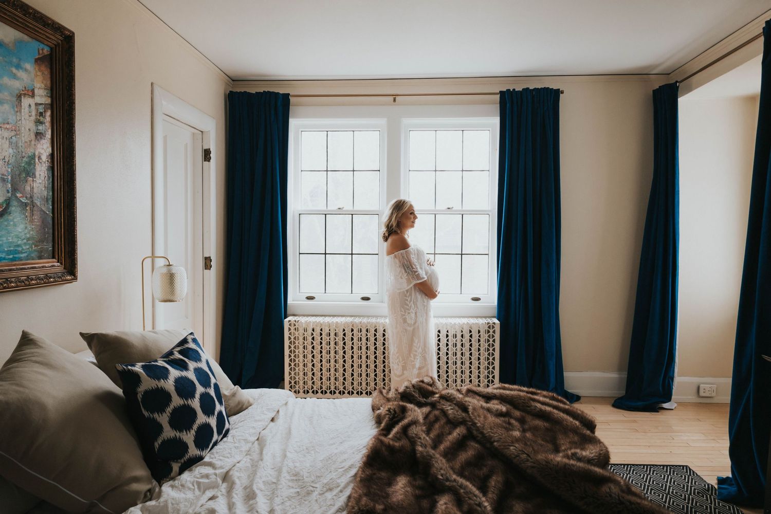 Silhouette of a woman in white standing by large windows with blue curtains in a bedroom.