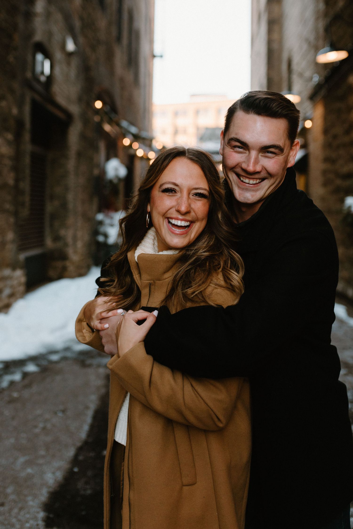 Couple embracing and laughing in snowy urban alley with string lights.