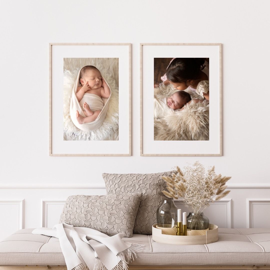 Two framed newborn photography portraits displayed above a decorative living room setup with neutral decor and dried flowers.