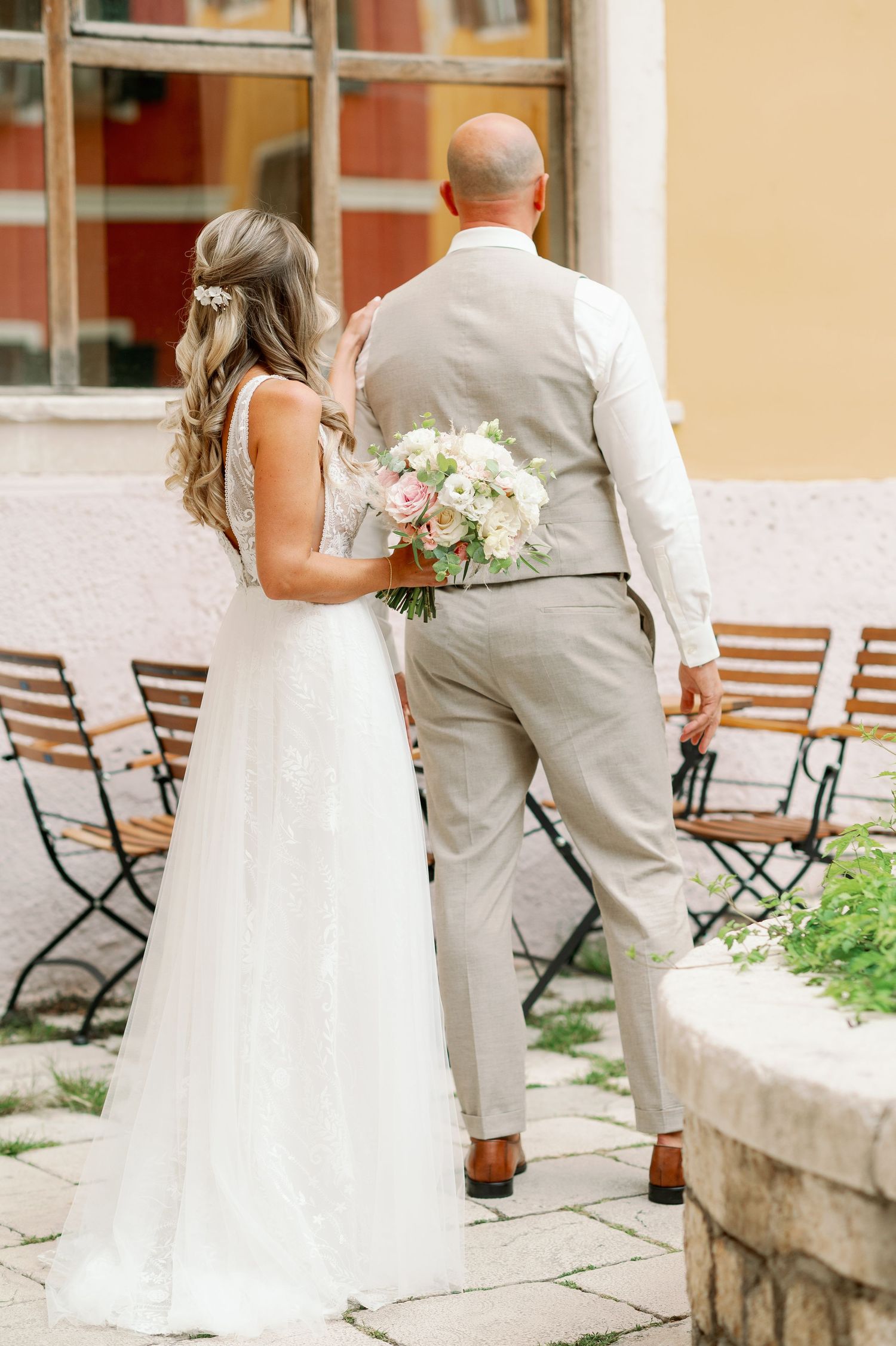 Bride and groom in light colored attire stand together on stone patio near folding chairs.