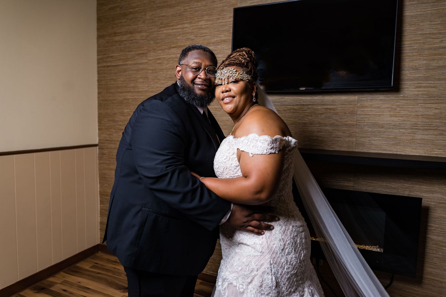 A romantic wedding couple embraces against a wooden wall with a mounted TV in an intimate indoor setting.