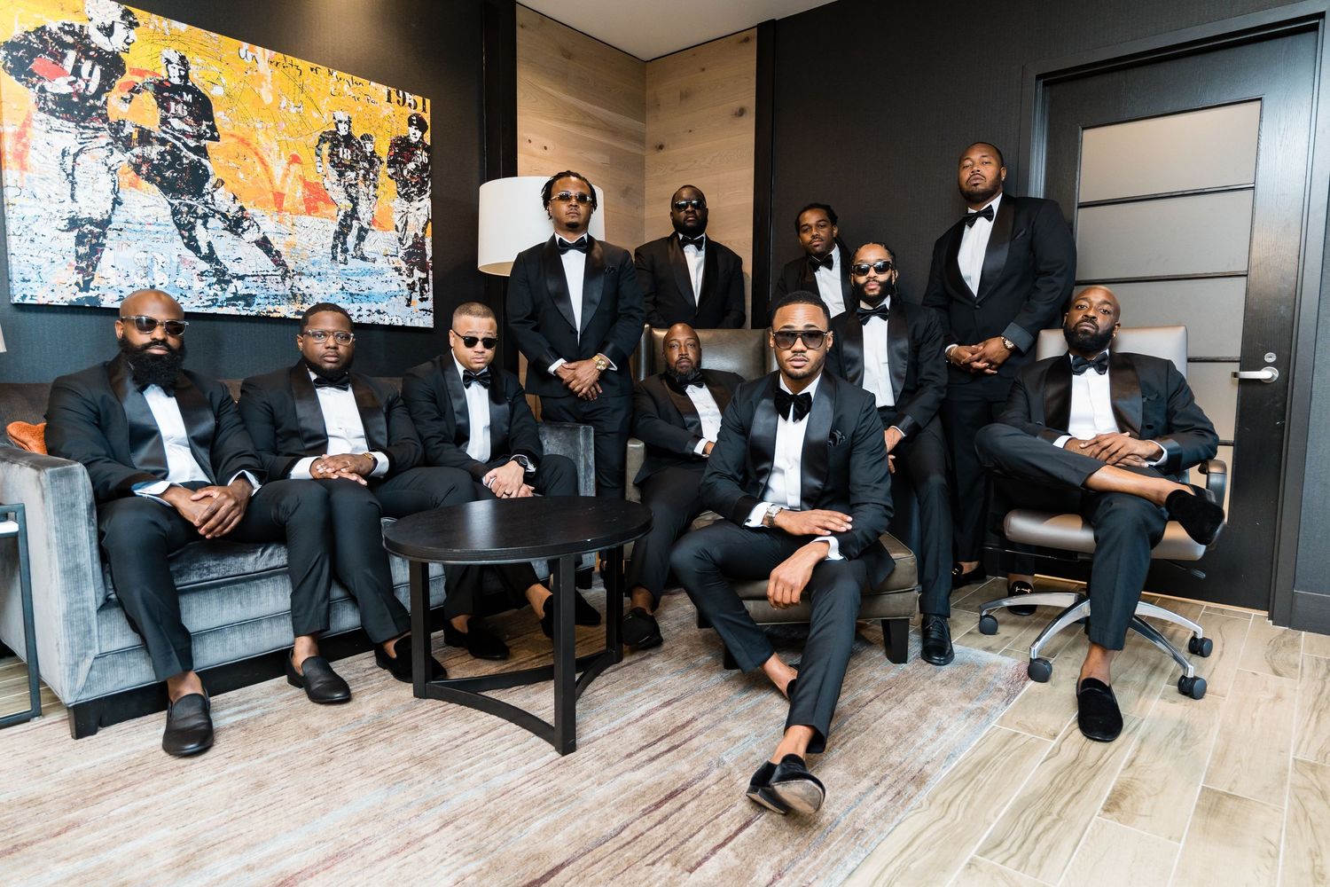 Groomsmen in black tuxedos gather in modern lounge setting for wedding photo.