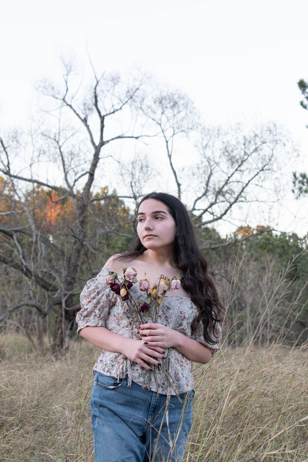 A romantic countryside portrait featuring a floral top and denim jeans among bare winter trees.