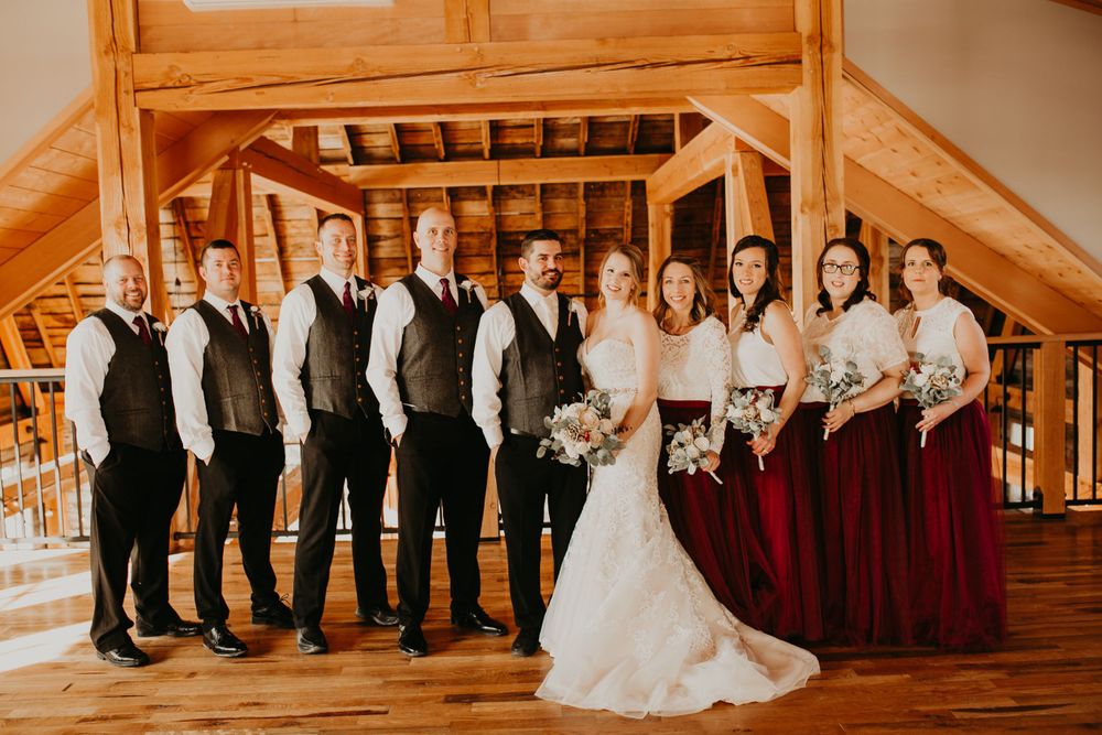 Wedding party in burgundy dresses and gray vests pose inside a warm rustic barn venue.