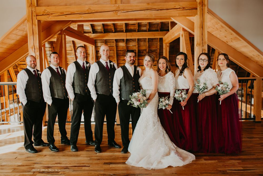 Bridal party with burgundy bridesmaids dresses and matching gray vests pose in timber barn.