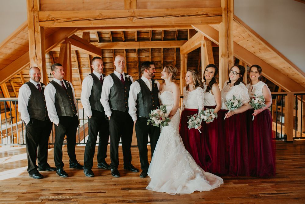 Wedding party poses in rustic barn, bridesmaids in burgundy gowns and groomsmen in gray vests.