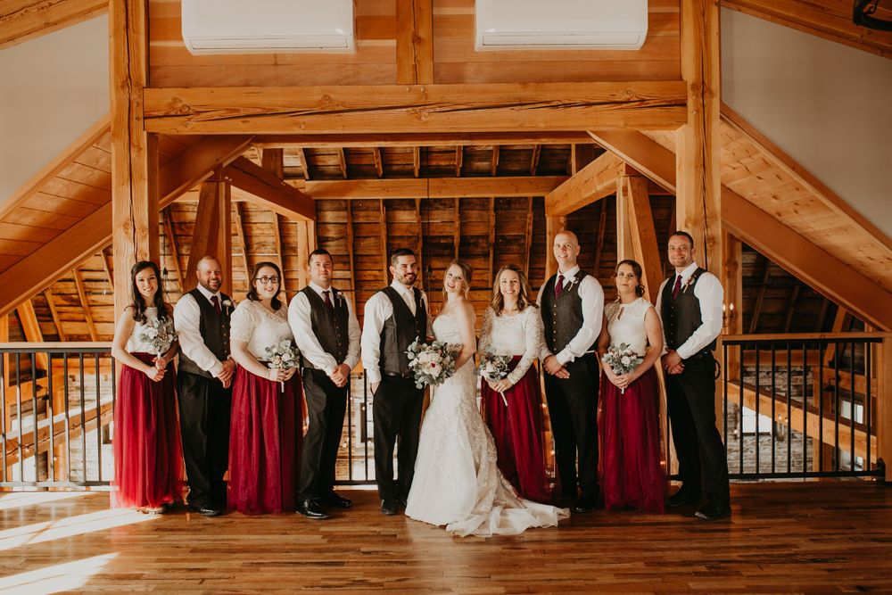 Full wedding party portrait inside a rustic wooden barn with exposed timber beam architecture.