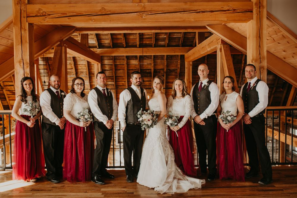 Bride and groom surrounded by wedding party in burgundy and gray inside a rustic barn venue.