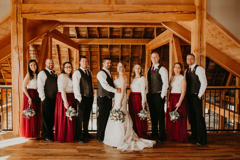 Bridal party smiles together in wooden barn, groomsmen in vests beside bridesmaids in red dresses.
